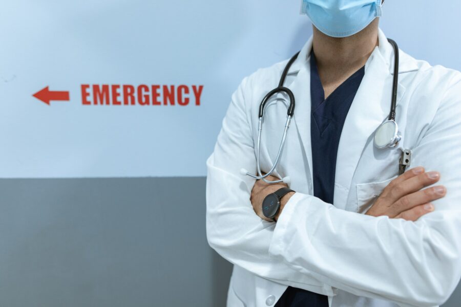 un medico a braccia incrociate sta in piedi davanti alla scritta "emergency". Indossa un camice bianco e al collo porta un fondendoscopio.