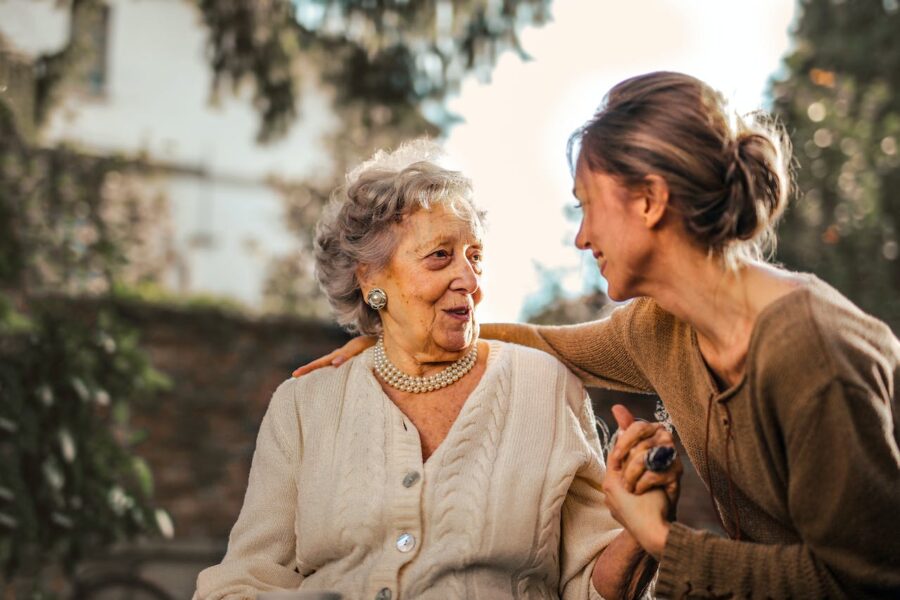 Assistenza domiciliare anziani. La foto mostra una signora anziana sorridente in compagnia di una donna più giovane. Le due donne sono sedute su una panchina in un parco.