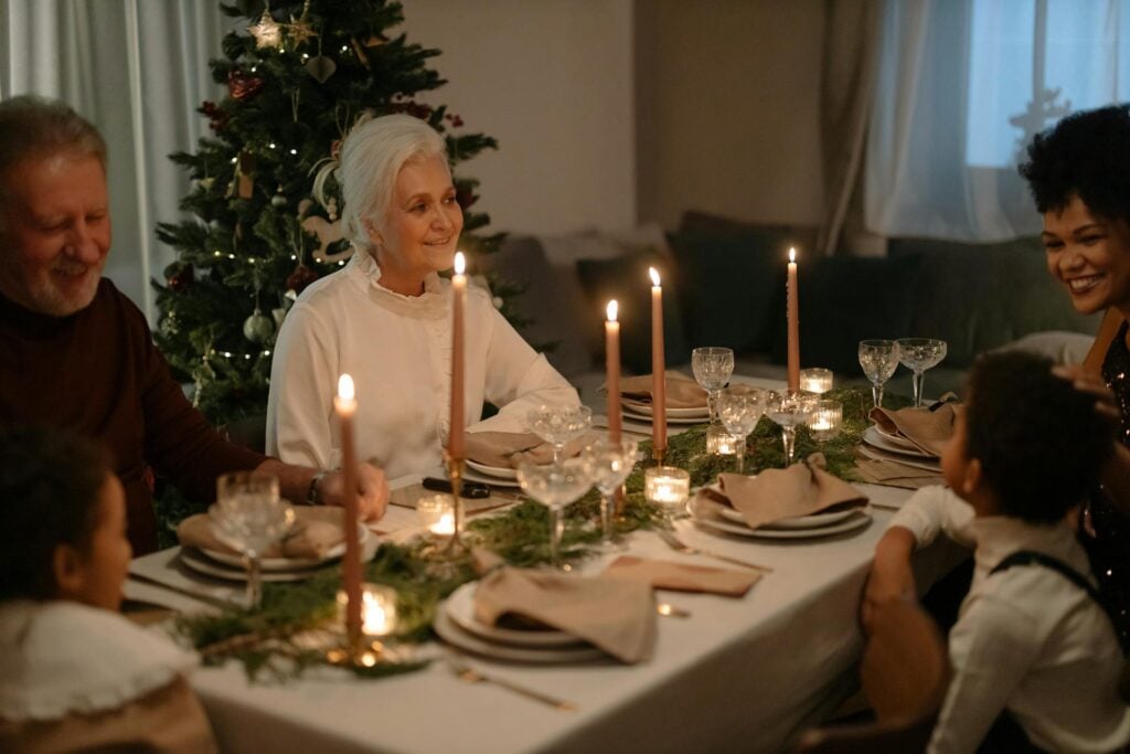isolamento sociale: stare loro vicino per abbattere la solitudine. Una signora anziana con i capelli raccolti si trova con altre persone seduta attorno a un tavolo decorato con candele a festeggiare il Natale. Sullo sfondo spicca un albero di Natale